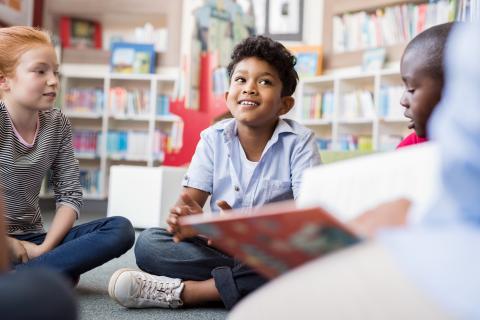 Several children smiling while holding books and sitting in the floor in the children's section of the library