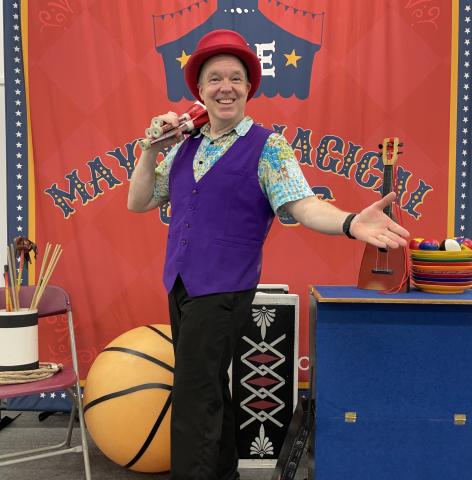 Gregory May from Circus Science poses in front of his 'Mayhew Magical Circus' backdrop holding juggling pins with his right hand and holding out his left hand in a welcoming gesture. Other Circus Science show props are in the background. 